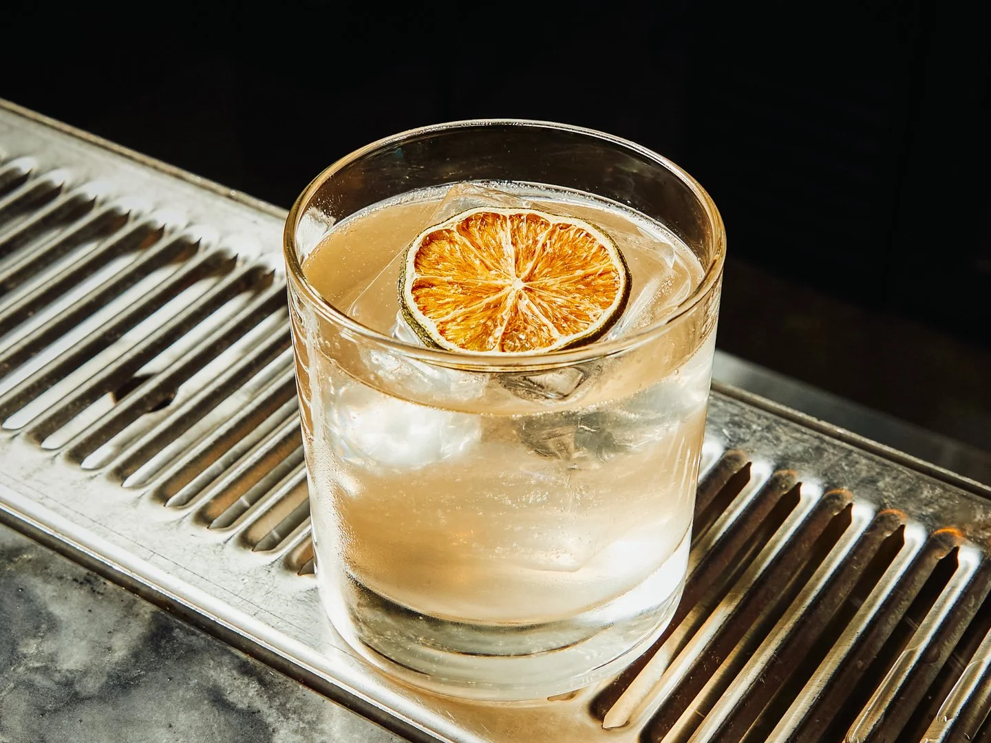 A glass of clear beverage with ice and a slice of dried orange on top, on a metal surface with slats.