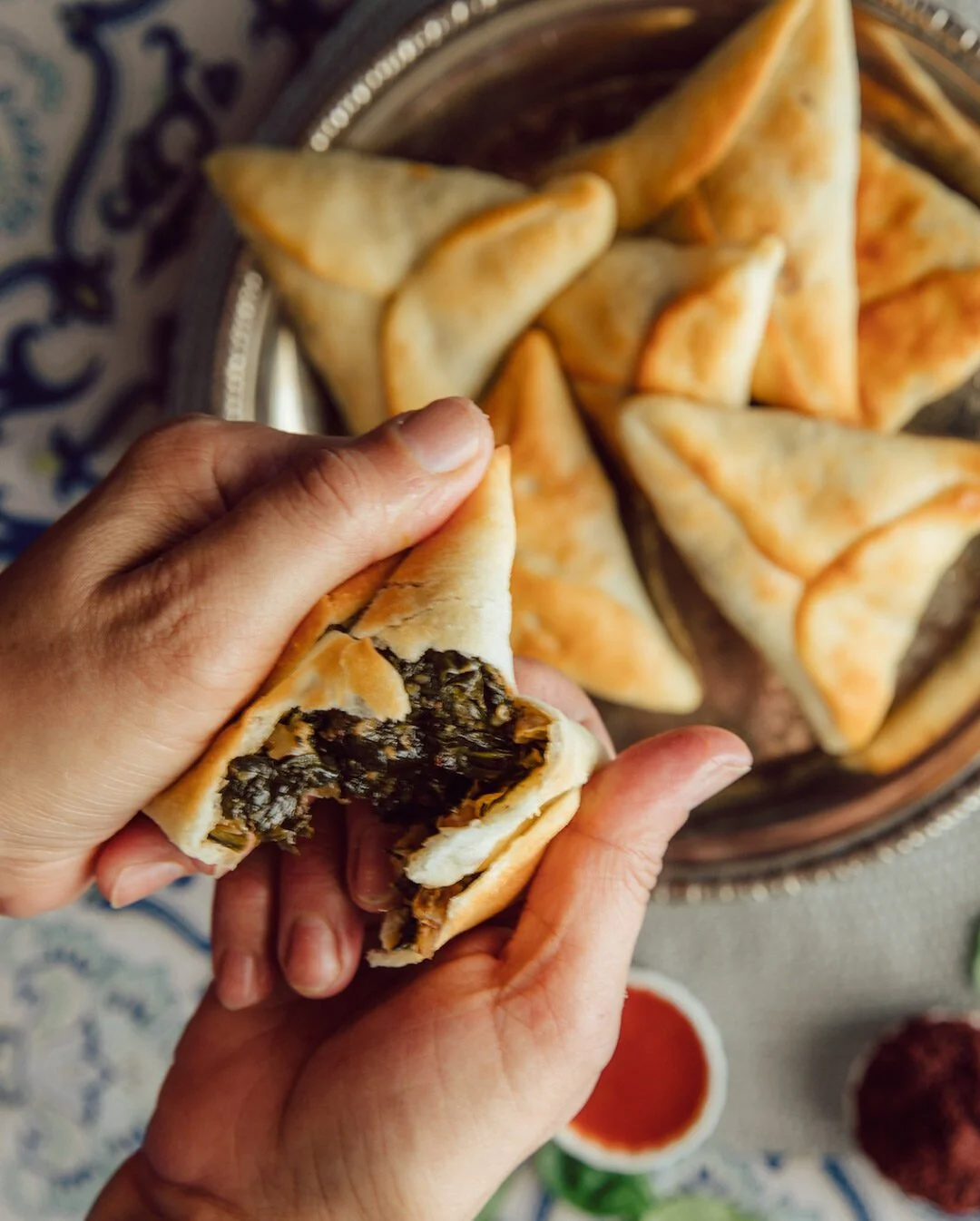 Person holding a partially eaten samosa filled with dark vegetable mixture, with a plate of more samosas and dipping sauces in the background.