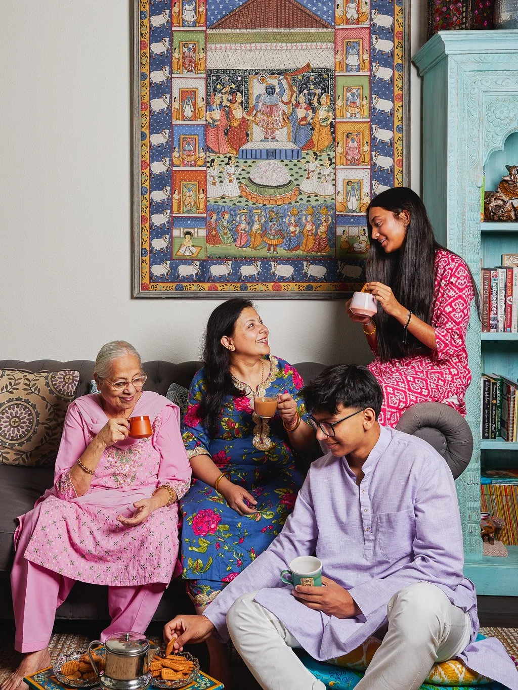Four women and one man enjoying tea and snacks in a cozy living room, with a colorful traditional Indian painting hanging on the wall behind them.