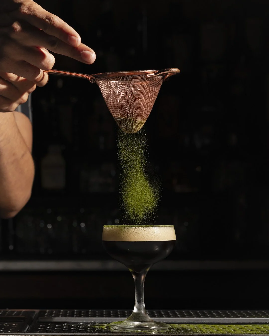 Green powder being sifted through a fine strainer into a black cocktail in a coupe glass.