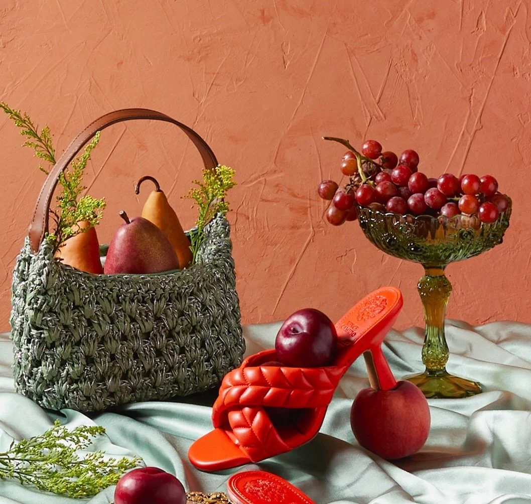 A woven basket with pears and a red apple, a red high-heeled shoe with another apple inside, a red apple on the fabric, and a glass pedestal bowl filled with red grapes, all set against an orange textured wall.