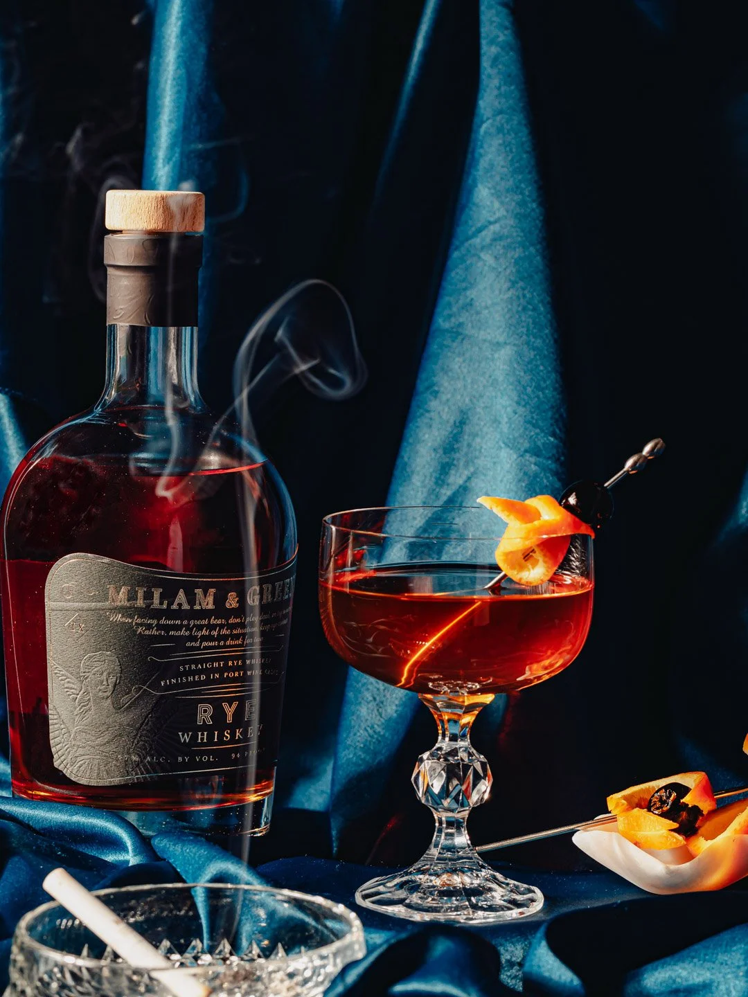 A bottle of rye whiskey, a cocktail glass with a red drink garnished with an orange peel, a small bowl with orange peel slices and garnish, and a glass bowl with a straw, all set against a dark blue satin background.