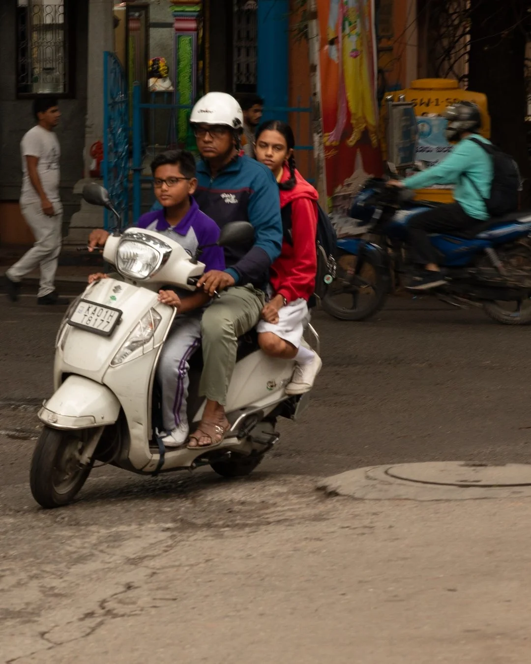 A man and two children riding a white scooter on a city street, with a busy background of pedestrians and other motorcyclists.