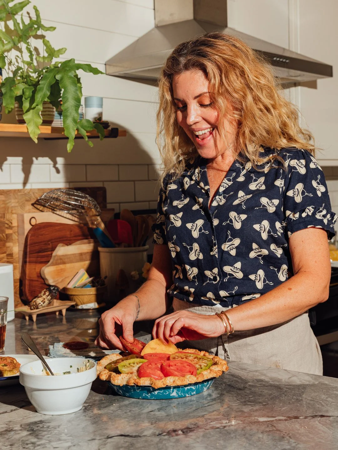 A woman with curly blonde hair smiling and preparing a vegetable tart with slices of tomato and other vegetables on a kitchen countertop.