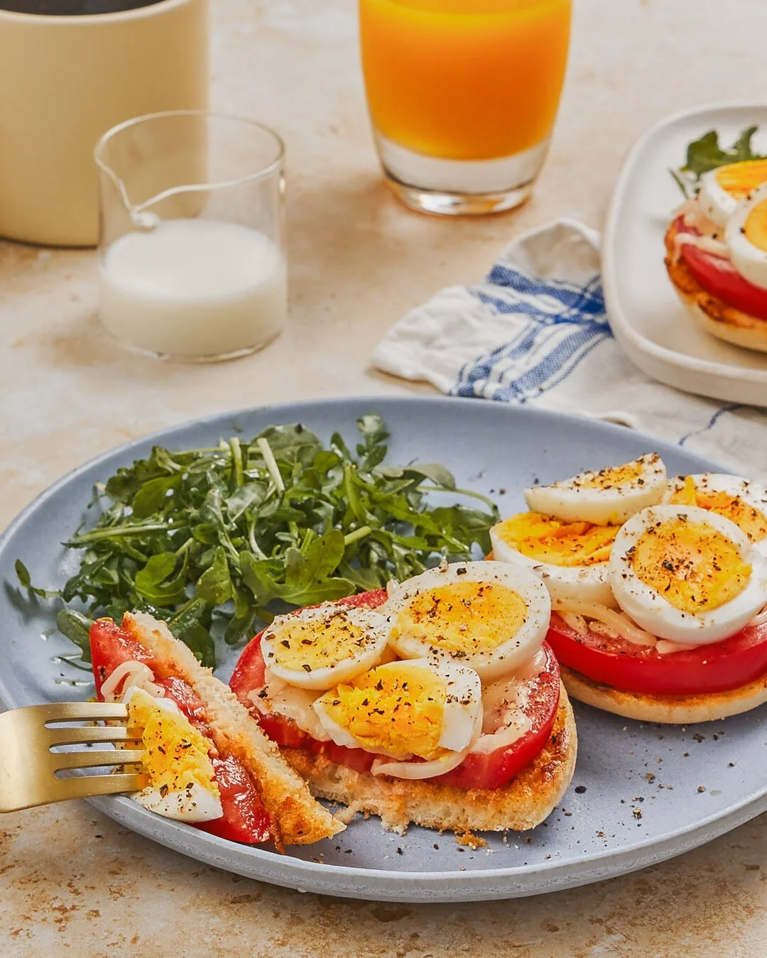 Plate with open-faced egg, tomato, and onion sandwiches with greens on the side, alongside a glass of orange juice, a small pitcher of milk, and a cup of coffee on the table.