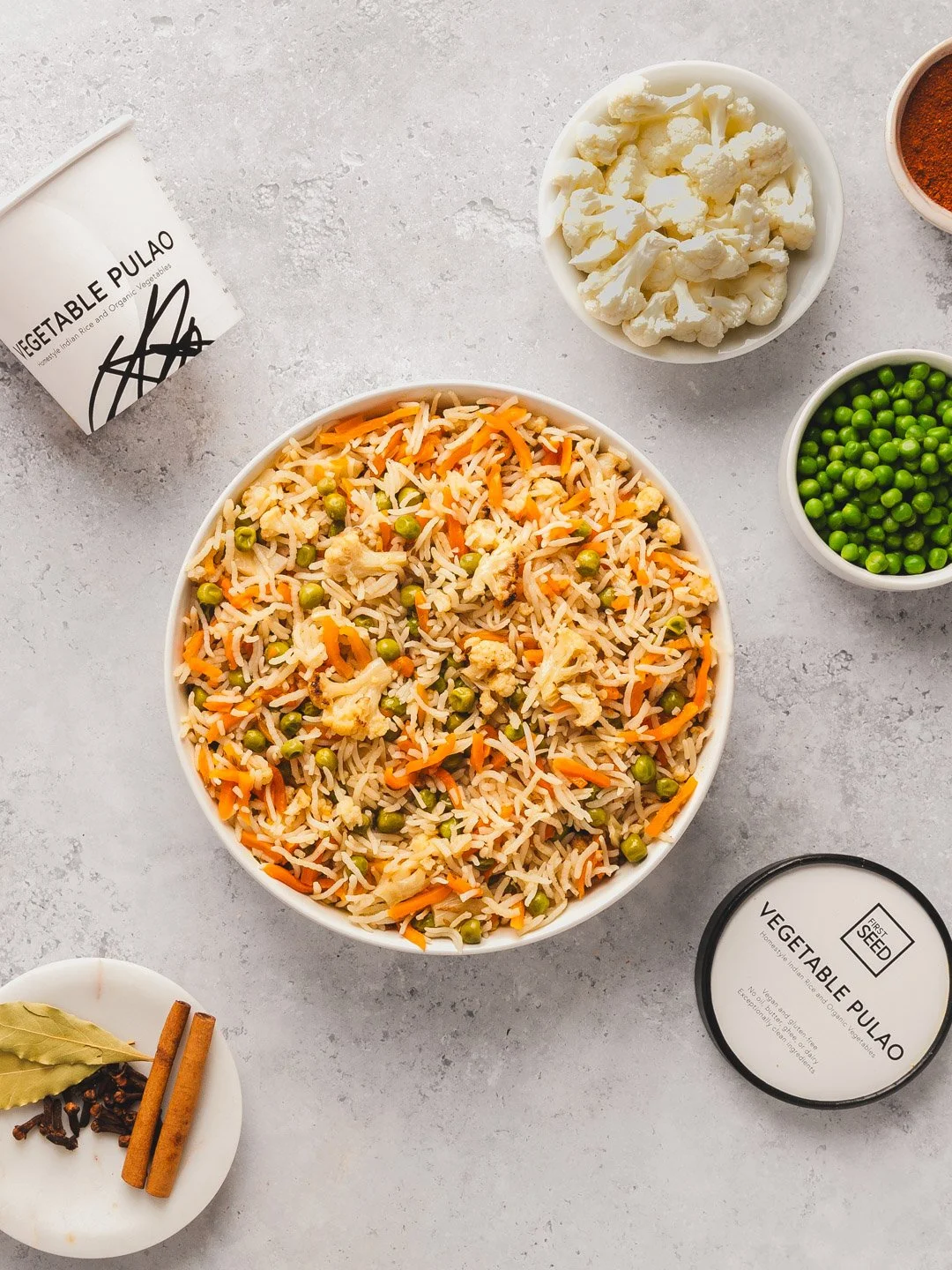 A bowl of vegetable pulao rice dish with peas, carrots, and cauliflower, surrounded by small bowls of cauliflower, green peas, and spices, with a container of vegetable pulao on the side.