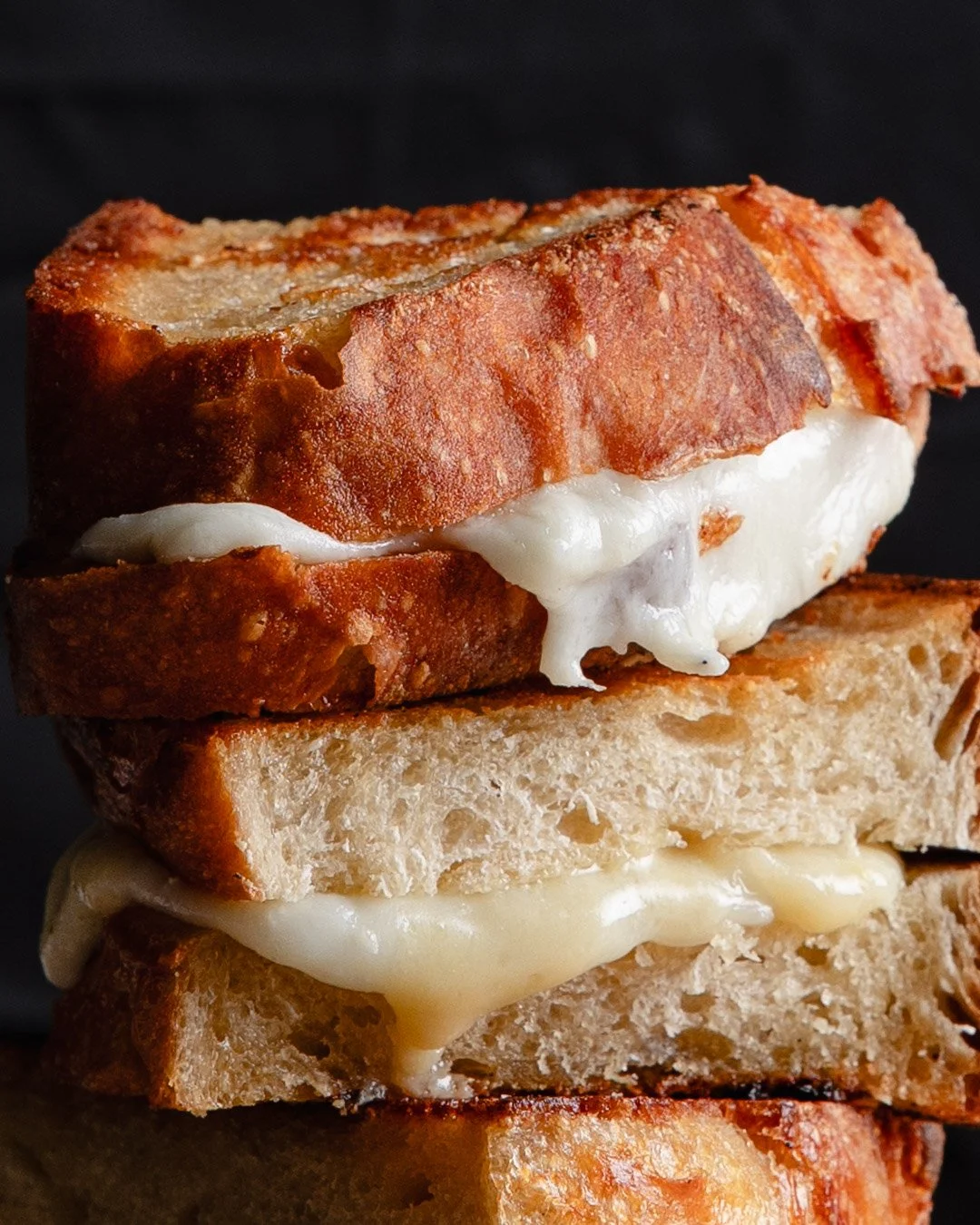 Close-up of a stacked bacon, bread, and melted cheese sandwich with crispy bacon on top and gooey cheese oozing from the slices on a dark background.