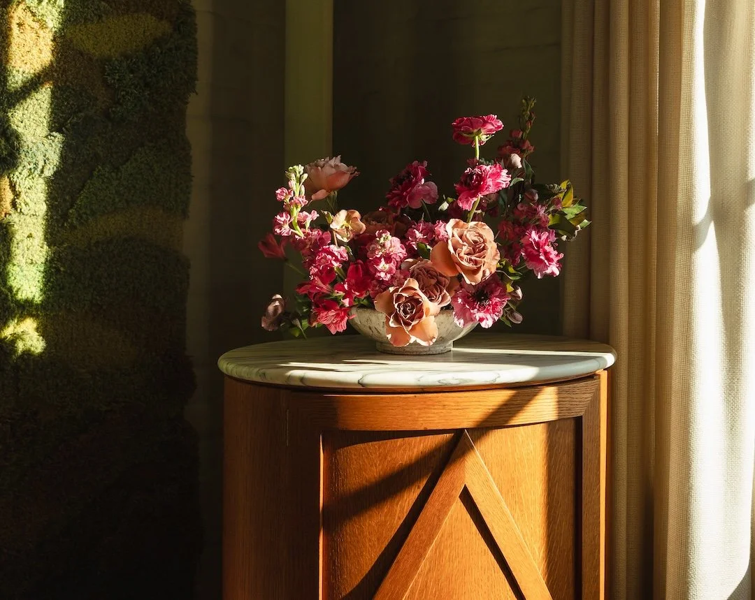 A white marble-topped wooden cabinet with a floral arrangement of pink and peach roses and pink flowers in a white vase, placed near a window with beige curtains, sunlight casting shadows on the wall and furniture.