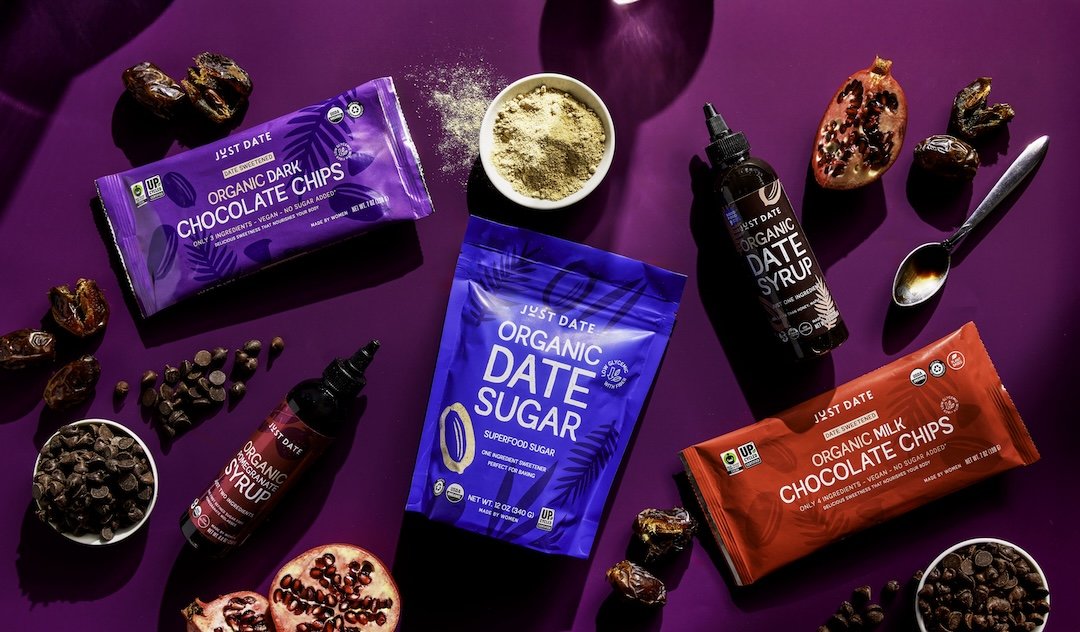 Various organic snack ingredients including chocolate chips, date sugar, date syrup, and a pomegranate, displayed on a purple background.