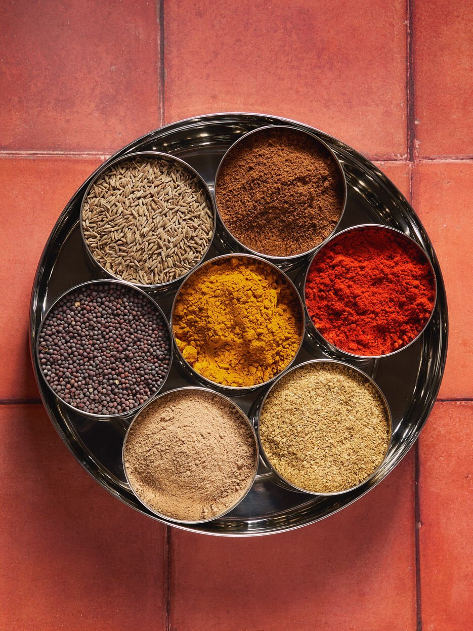 Metal tray with seven bowls filled with various spices and herbs, placed on a red tiled floor.