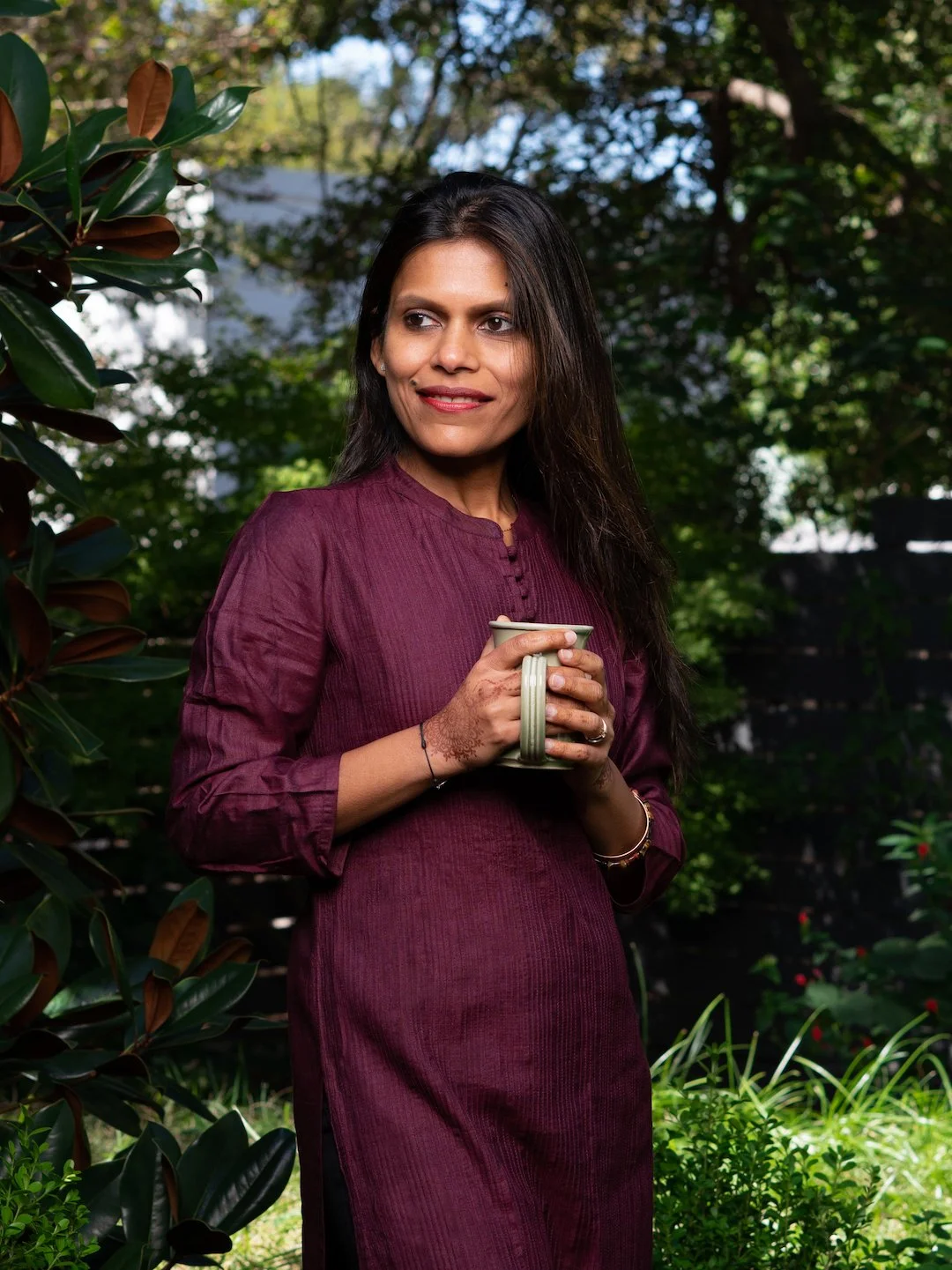 A woman with long dark hair wearing a maroon dress holding a beige coffee mug outside in a garden with green plants and trees.
