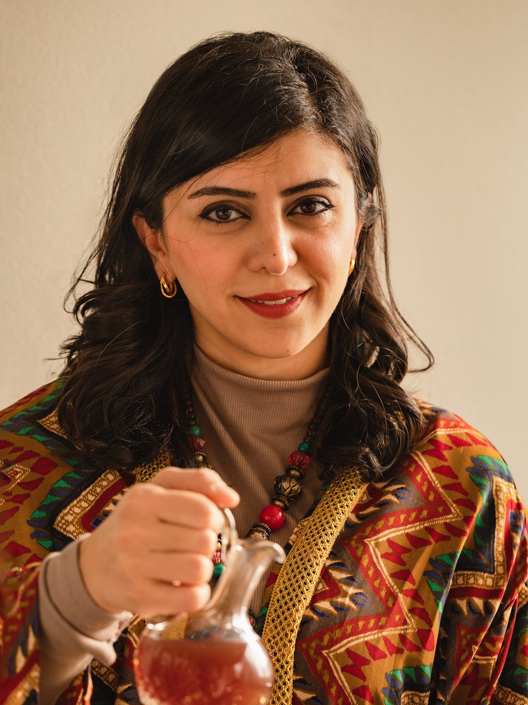 A woman with dark hair and a warm smile, wearing colorful, patterned clothing and jewelry, holding a small glass pitcher.