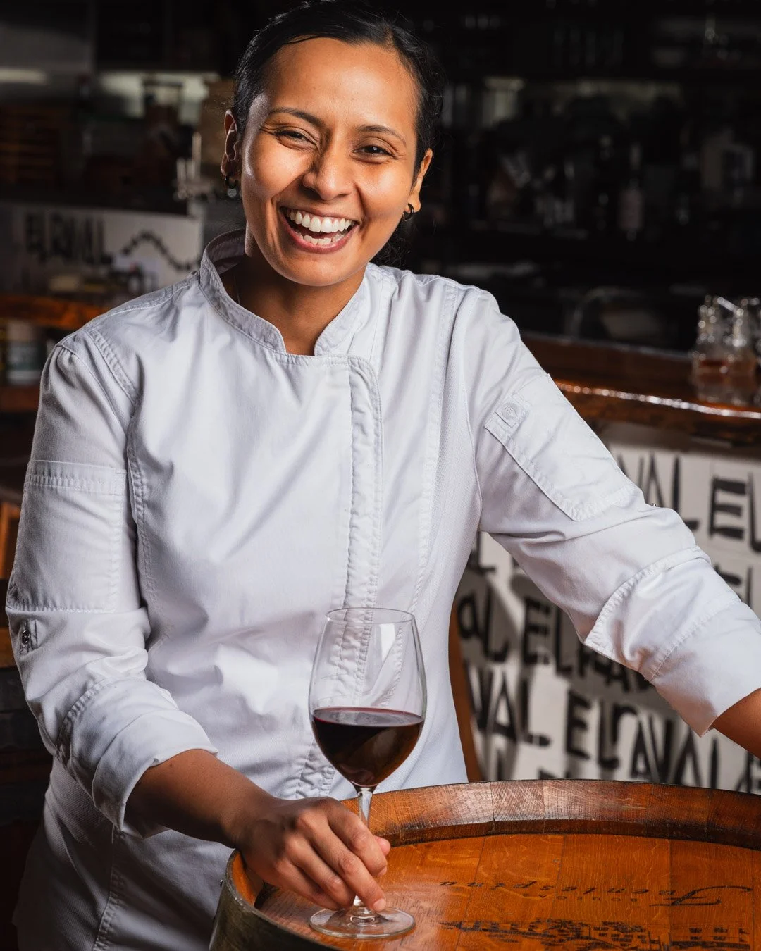 Smiling woman in white chef's coat holding a glass of red wine in a bar or restaurant setting.