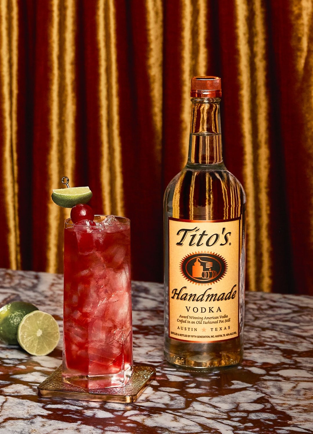 A bottle of Tito's Handmade Vodka on a marble table next to a glass of pink cocktail with ice, garnished with a lime wedge and cherry, with a lime half in the background. Gold curtains are in the background.