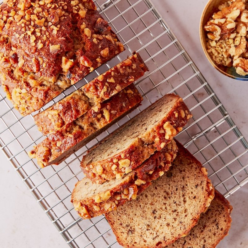 Banana bread with chopped nuts on top, sliced, cooling on a wire rack with a bowl of chopped nuts nearby.