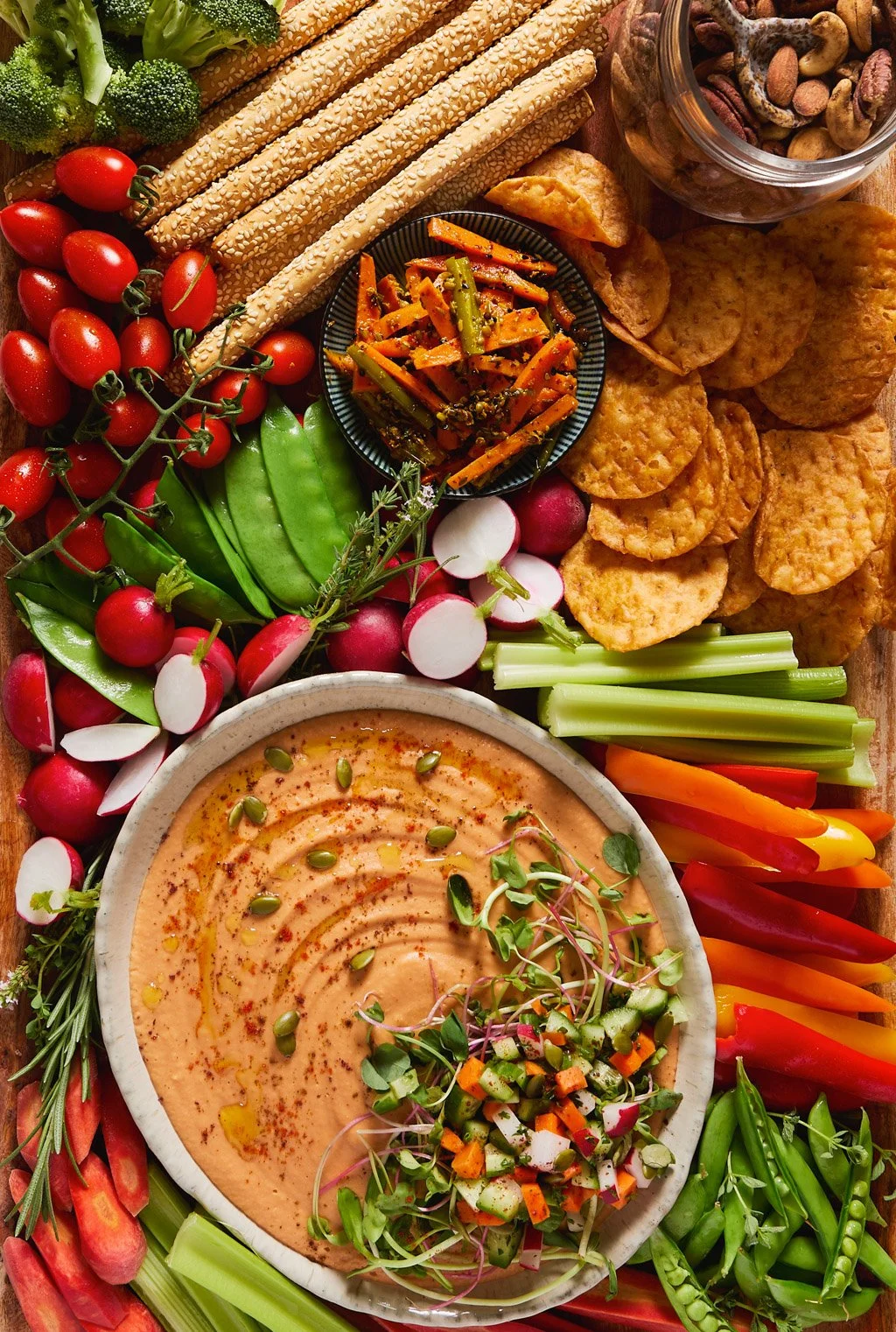 Colorful vegetable platter with cherry tomatoes, radishes, celery, carrots, snap peas, and broccoli, accompanied by hummus, sesame sticks, crackers, and a bowl of mixed nuts.