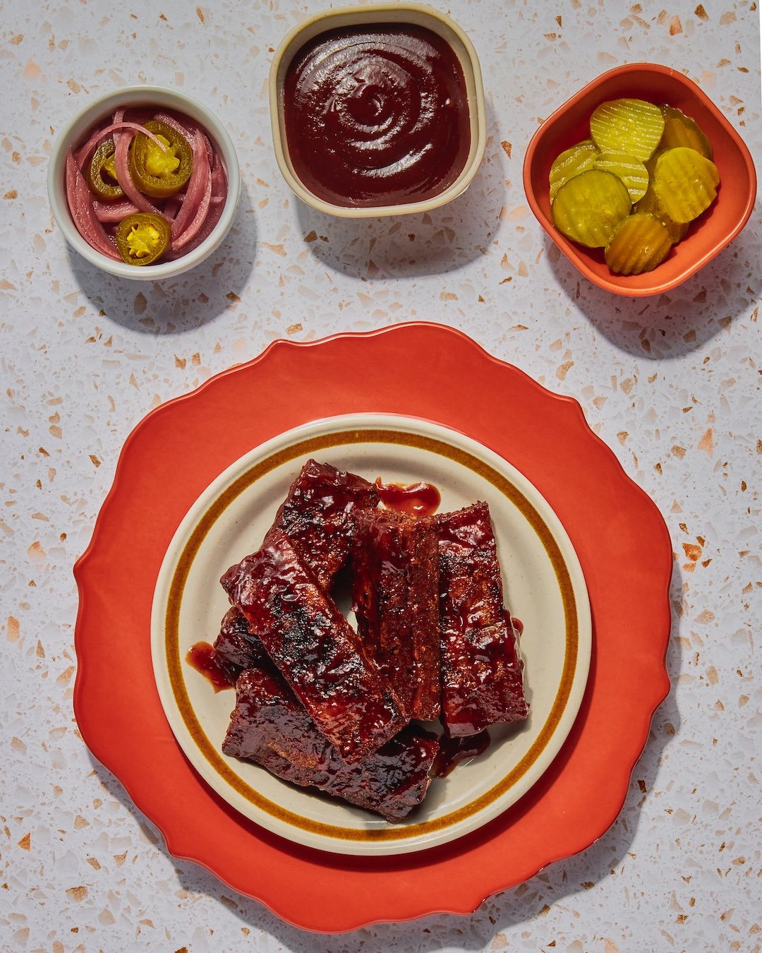 Barbecue ribs on a decorative plate with three small bowls of condiments and pickles above it.