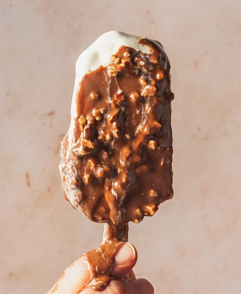 Hand holding a dessert popsicle covered in chocolate with nuts, partially coated in vanilla ice cream.