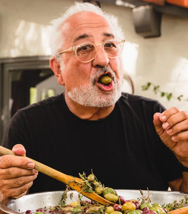 Older man with white hair, beard, glasses, wearing a black shirt, holding a wooden spoon, enjoying olives from a large bowl.