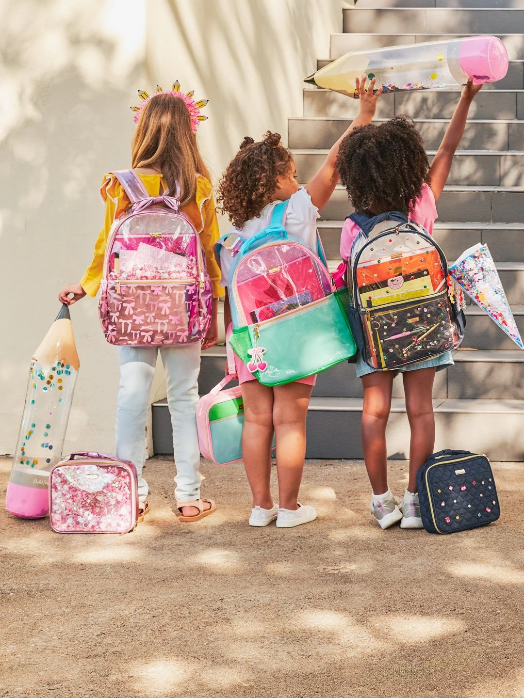 Four young girls with backpacks standing on stairs, holding oversized bubblegum pouches and pushing large pencil-shaped bubbles.