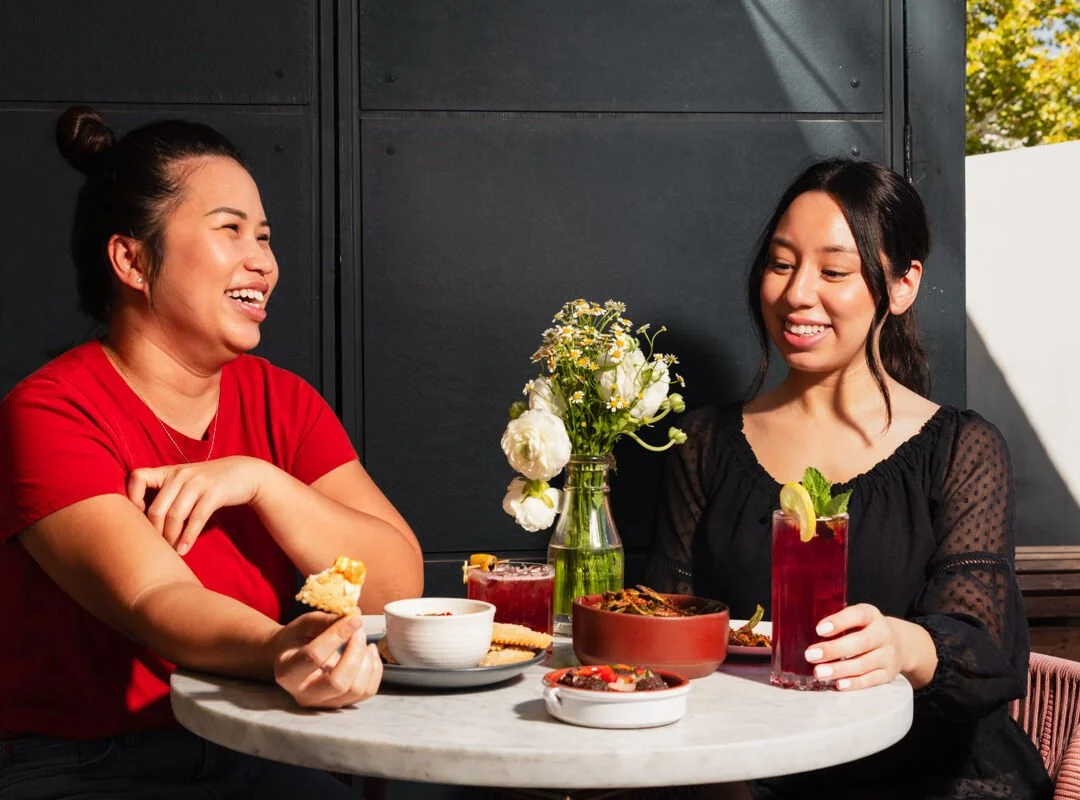 Two women laughing and enjoying drinks at an outdoor table with food and a vase with flowers.