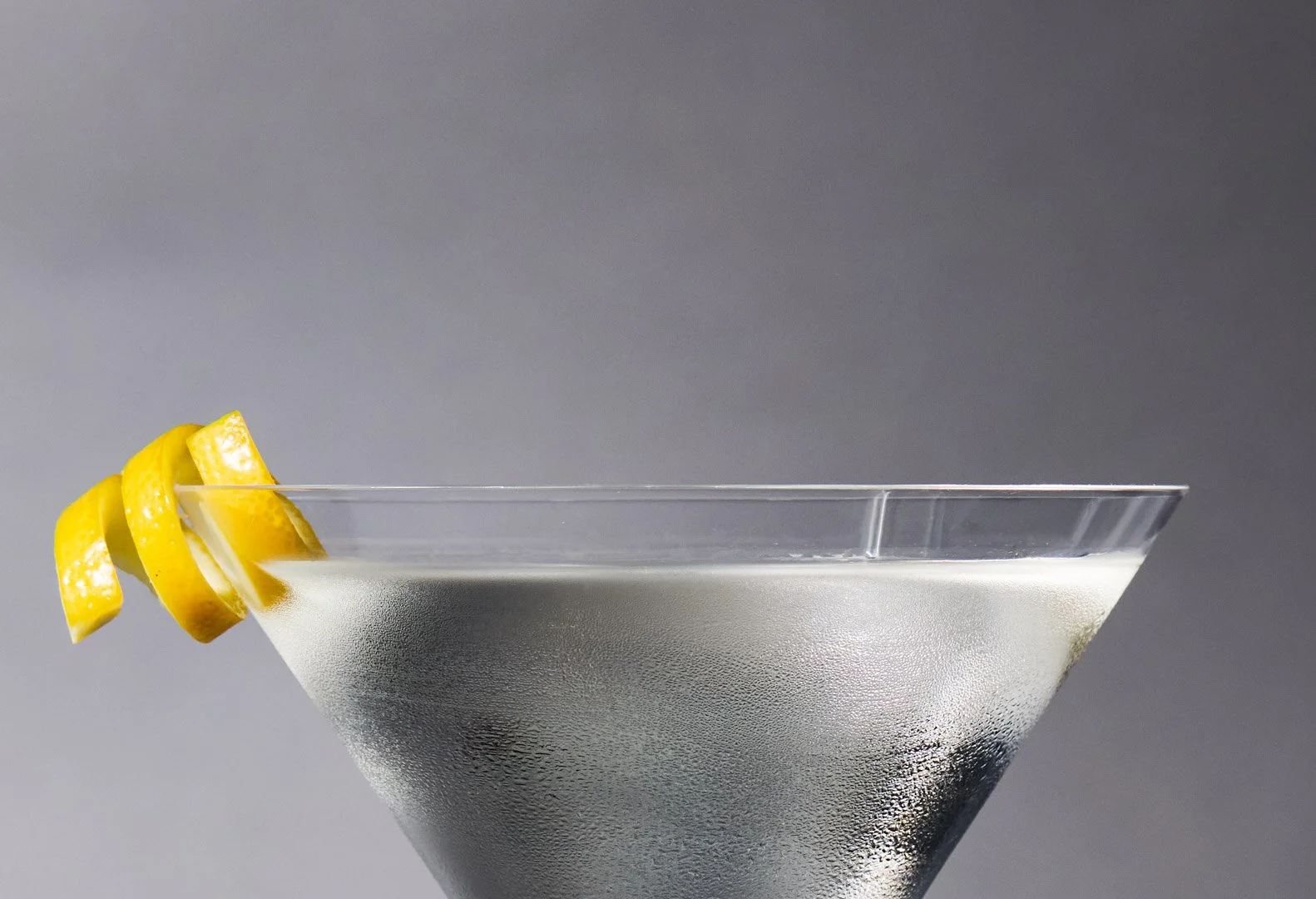 A martini glass filled with a clear liquid, garnished with three lemon twists on the rim, against a plain gray background.
