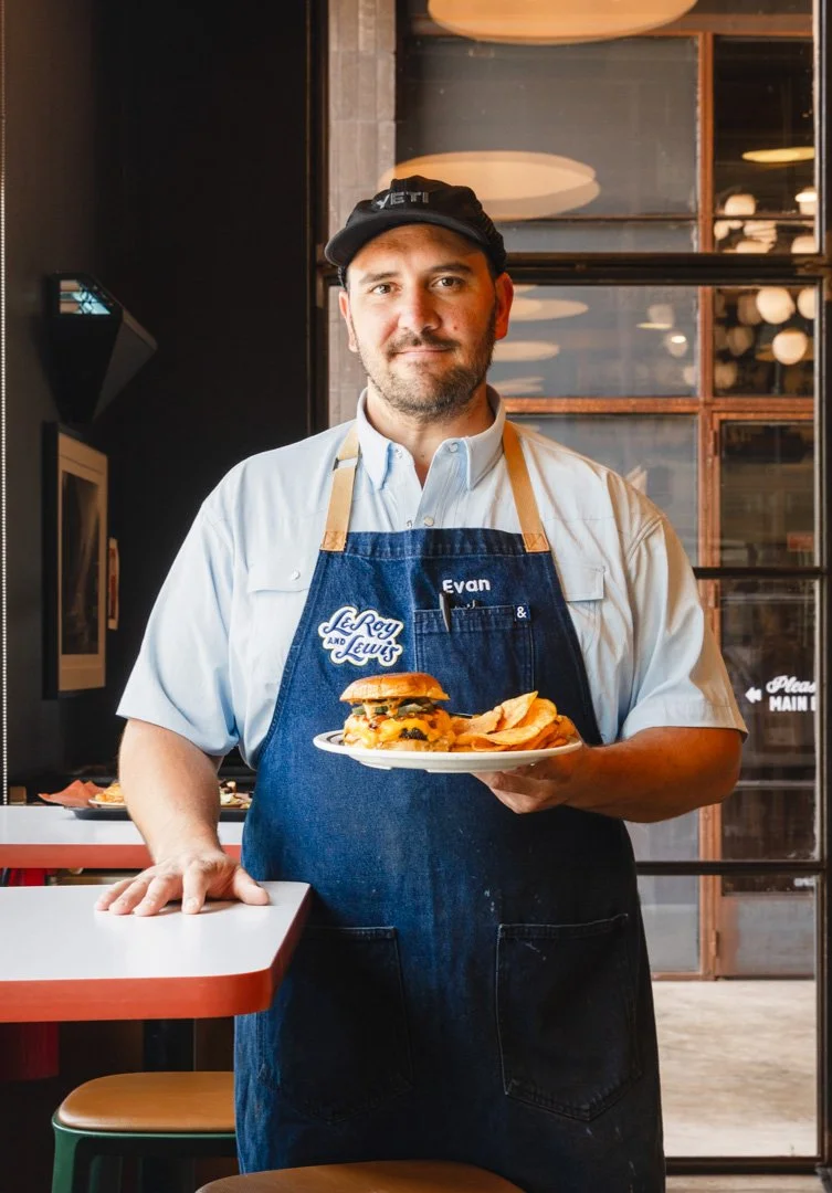 A man wearing a chef's uniform and apron, holding a plate with a burger and potato chips inside a restaurant.