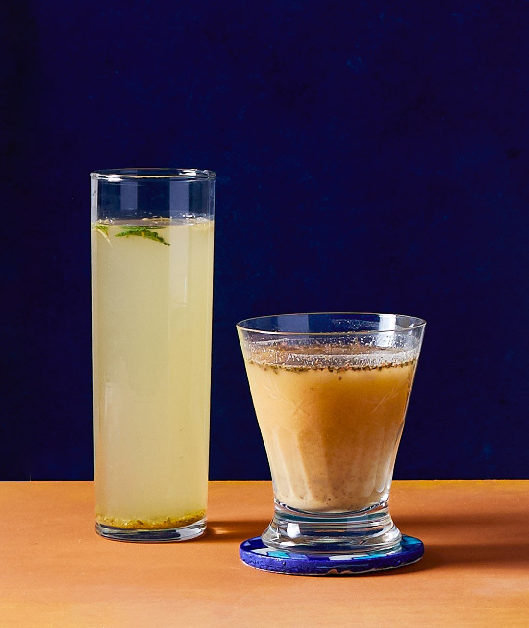 Two drinks on a wooden surface: a tall glass of pale yellow lemonade with floating lime wedges and a short glass of frothy iced coffee or tea.