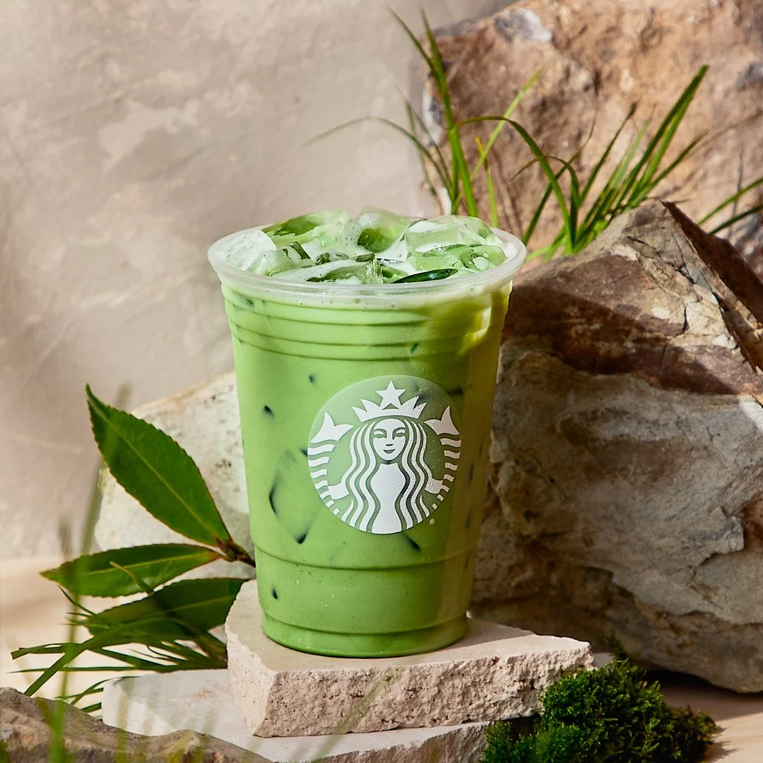 A Starbucks iced matcha latte with ice cubes in a clear plastic cup, set on a stone and surrounded by rocks and green plants.