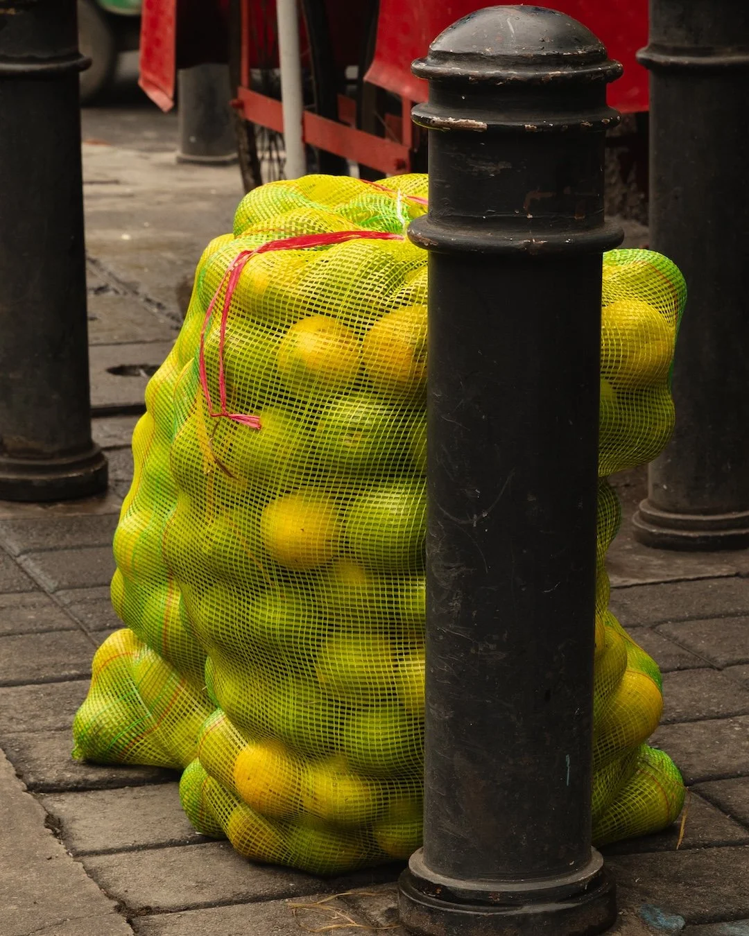 A mesh bag filled with yellow and green apples is tied with red string and resting against black metal posts on a city sidewalk.