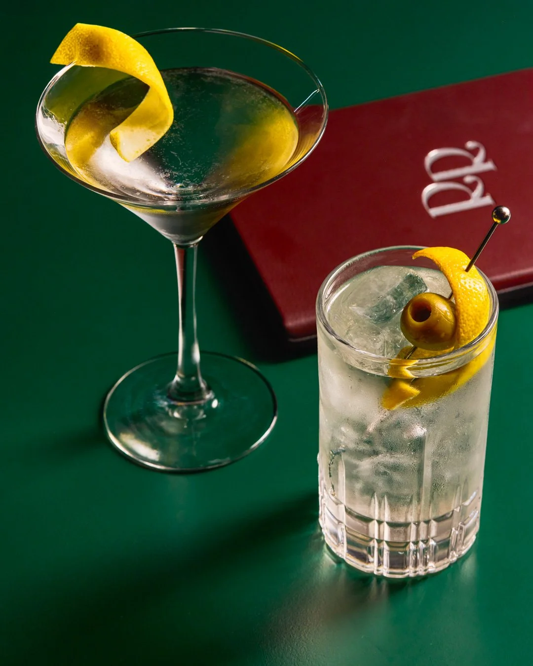 Two cocktails with lemon garnishes, one in a martini glass and one in a highball glass with ice and an olive. A red menu or book is in the background.