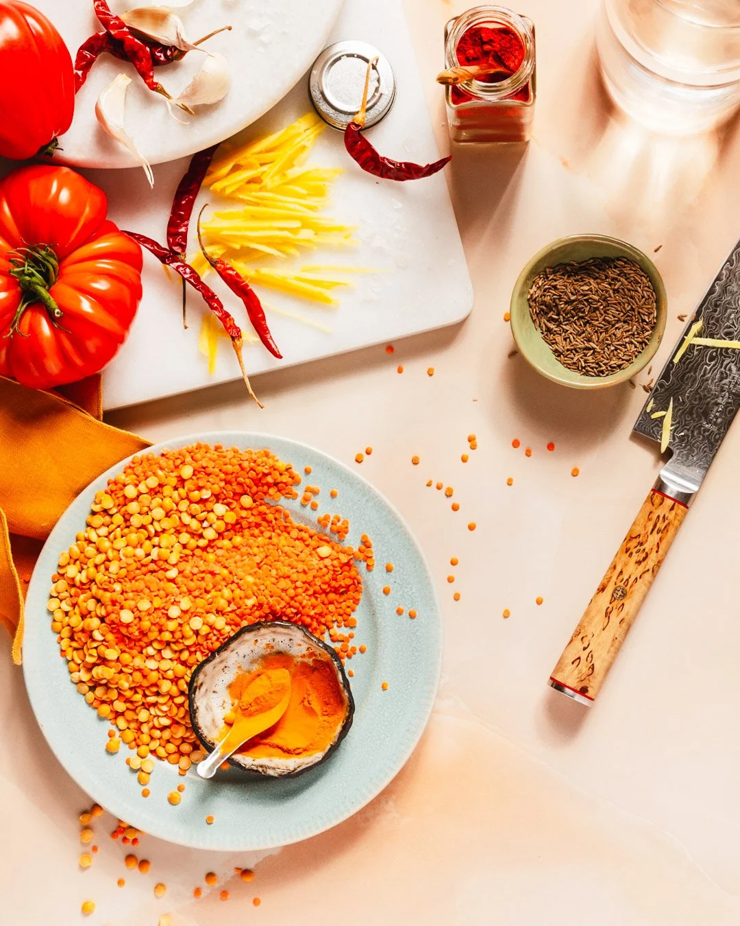 Assorted ingredients for cooking, including red tomatoes, garlic, dried red chili peppers, shredded cheese, red spice, cumin seeds, yellow lentils, turmeric in a halved coconut shell, a kitchen knife, and a light-colored marble surface.