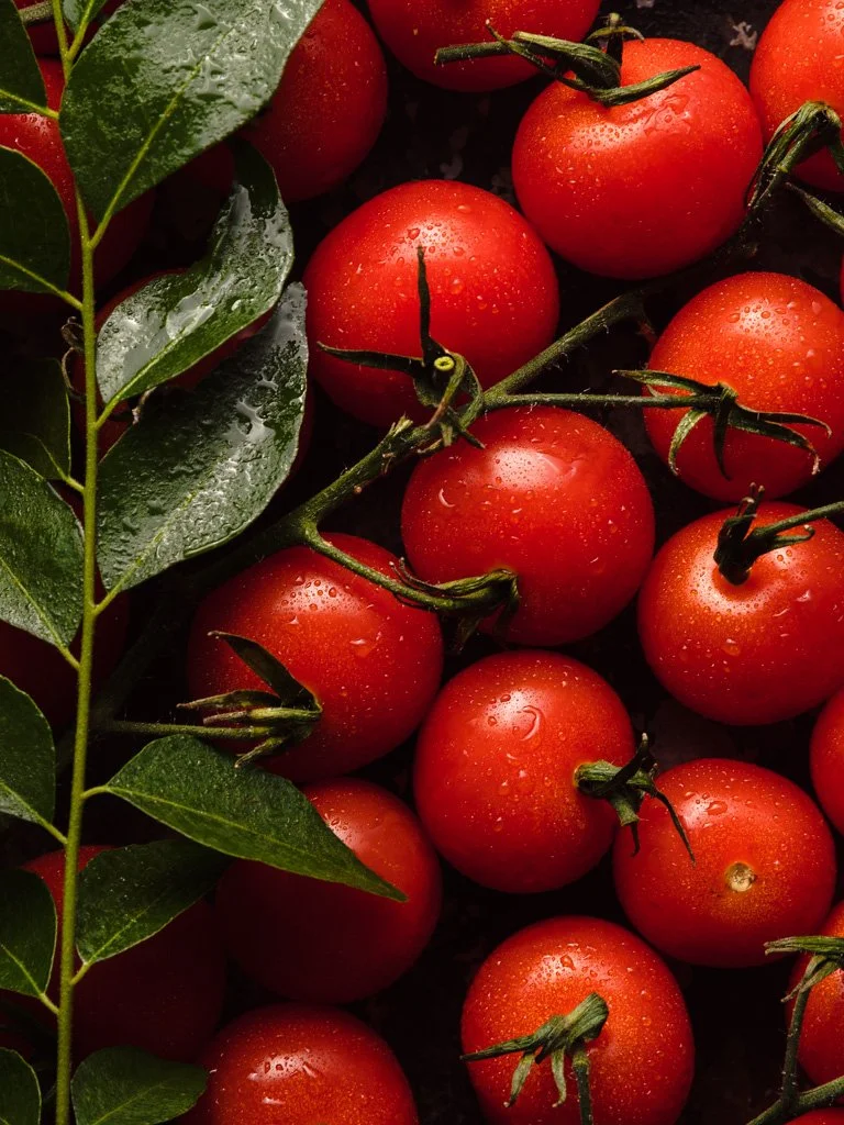 Cluster of ripe red cherry tomatoes with green stems and water droplets, alongside dark green leaves with water droplets.