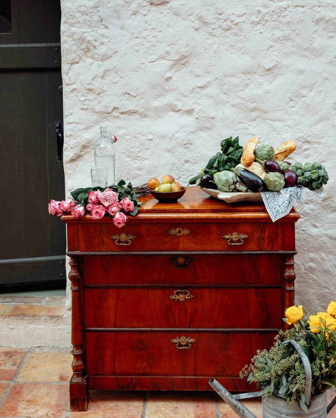 An antique wooden dresser with a bouquet of pink roses, a bowl of apples, and a fruit and vegetable display on top. A glass water bottle and a vase of yellow flowers are also nearby. The background features a rustic white wall and a tiled floor.