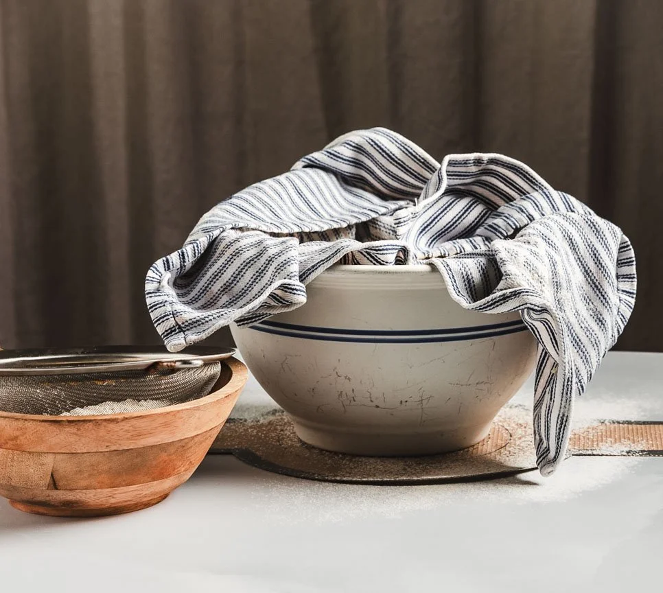 A ceramic bowl with a striped cloth draped over it, placed on a table with a wooden bowl and a metal sieve on the side.