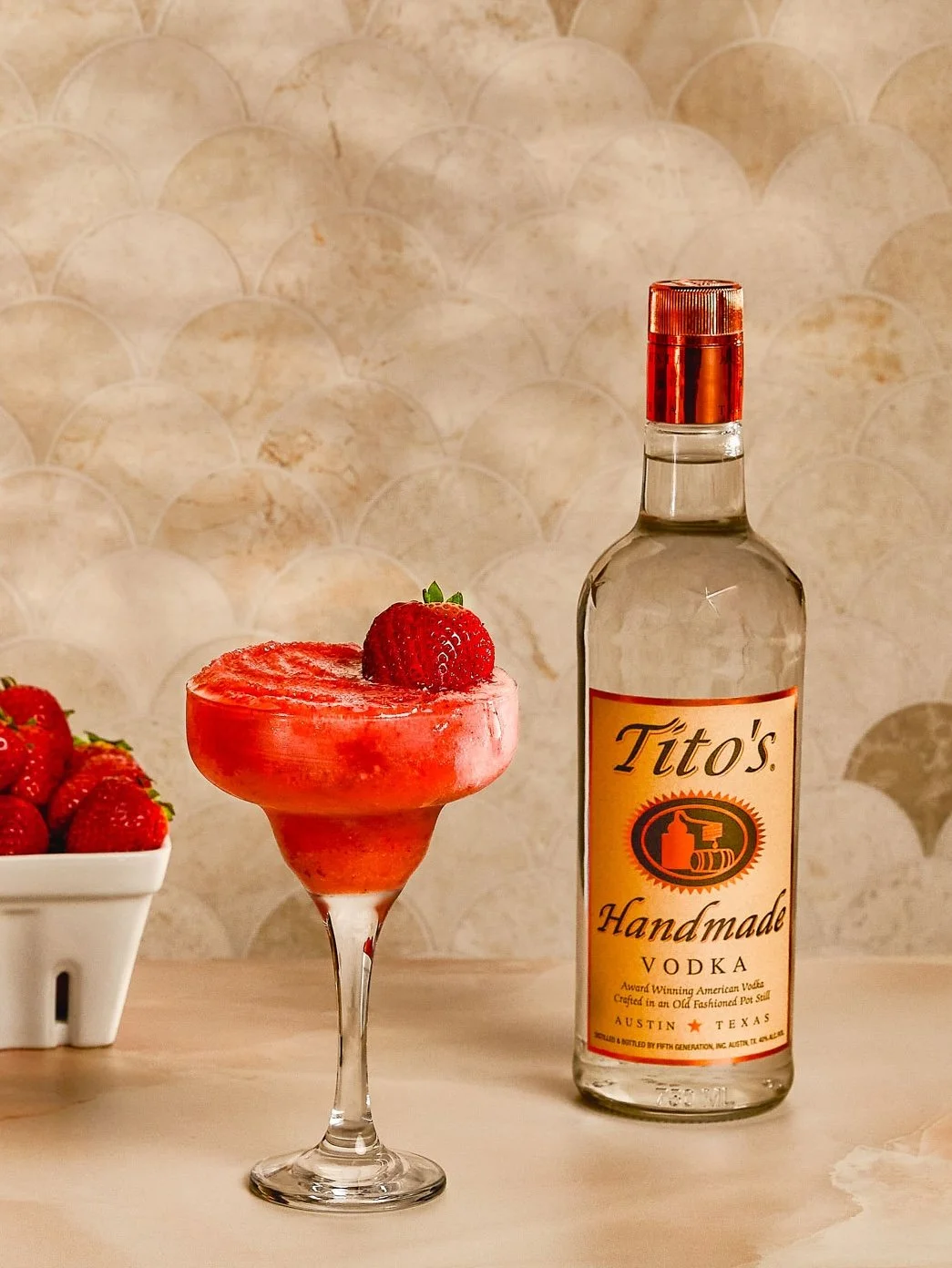 A strawberry daiquiri cocktail garnished with a strawberry on top of the glass and a bottle of Tito's handmade vodka next to it, with a bowl of strawberries in the background.