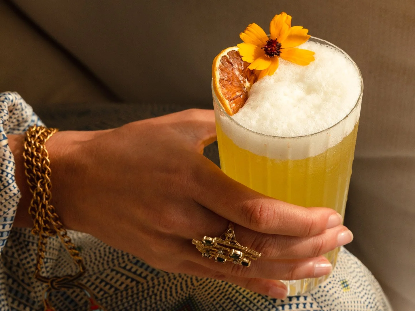 A hand holding a tall glass of yellow beverage with a frothy top, garnished with a slice of dried orange and a yellow flower.