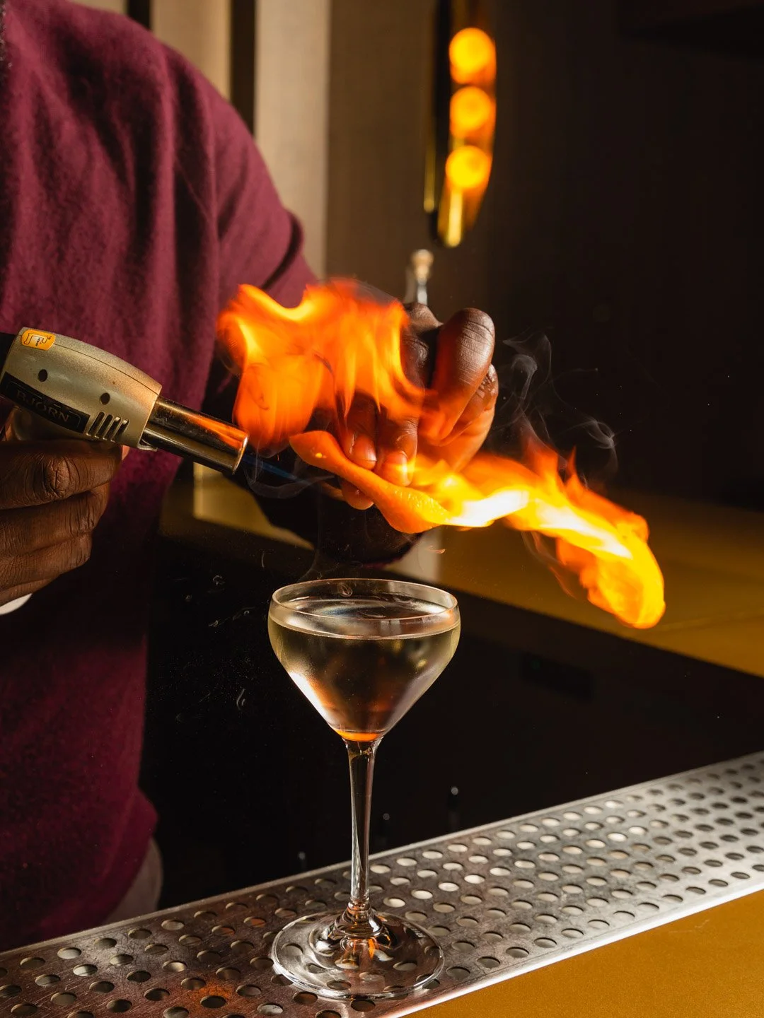 Bartender using a torch to ignite a flaming orange peel over a cocktail in a martini glass.
