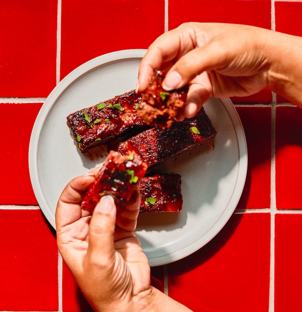 Hands holding two pieces of glazed barbecue ribs with green garnish over a white plate on a red tiled surface.