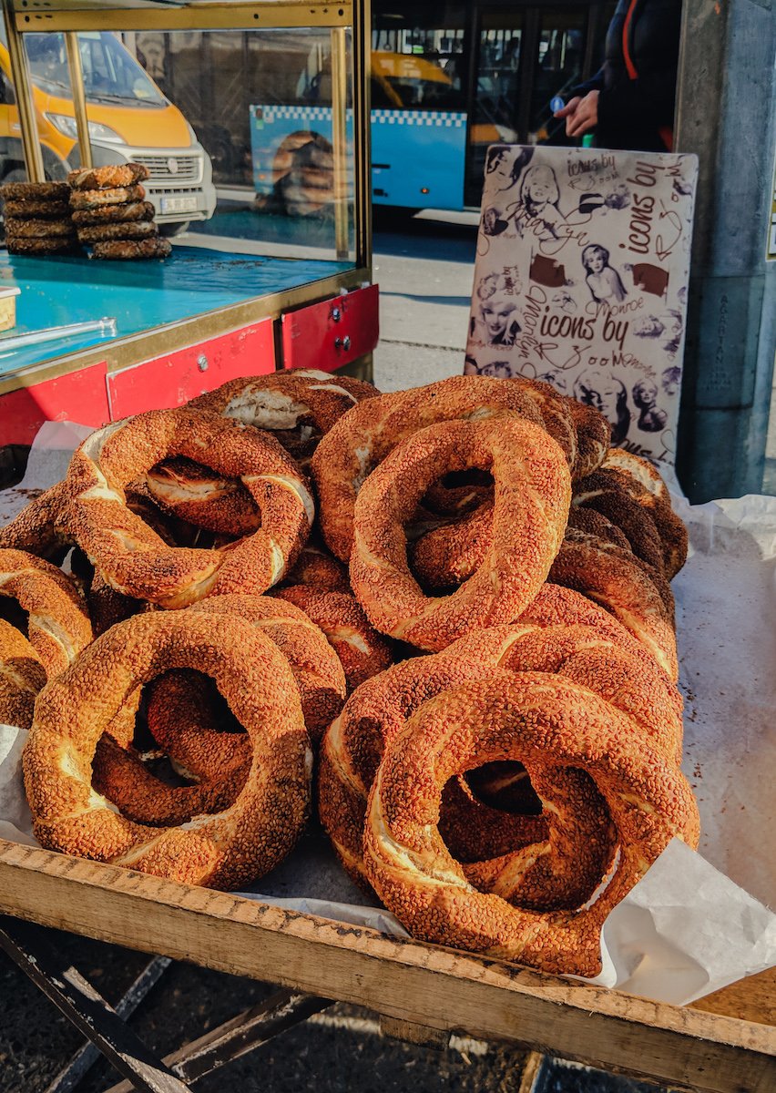 A tray of freshly baked sesame seed bagels on a street vendor cart, with some in a display case and others on parchment paper, surrounded by cars and a bus in the background.