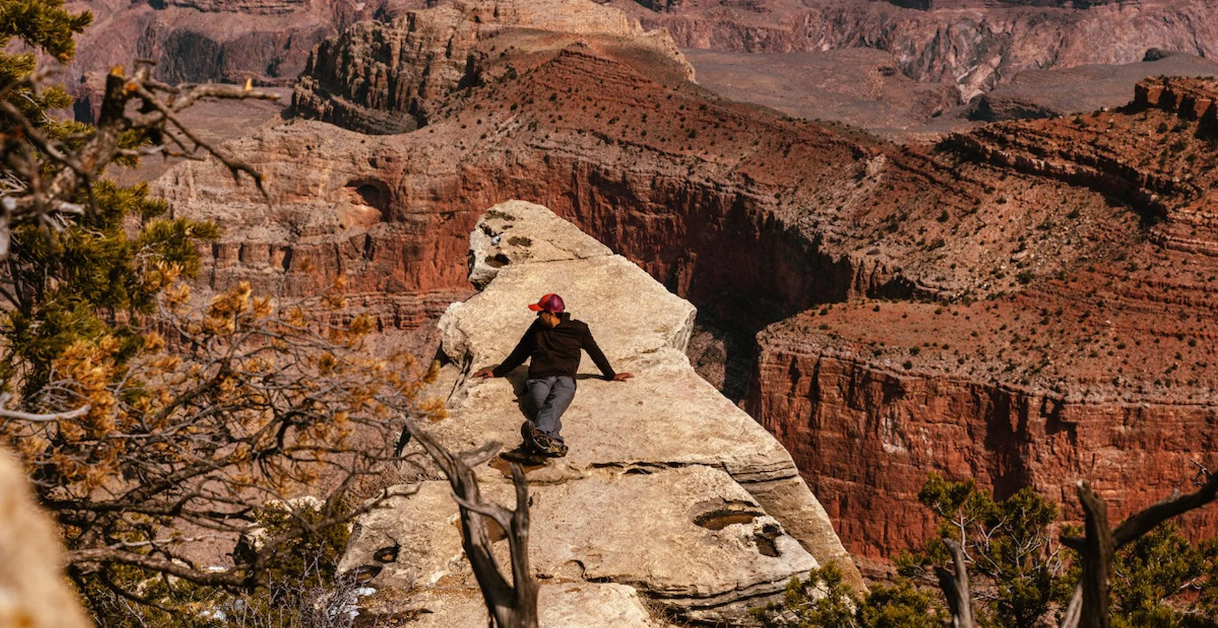 A person wearing a black jacket, gray pants, and a red cap rocks and balances on the edge of a large sandstone formation overlooking the Grand Canyon with layered red rocks and cliffs in the background.