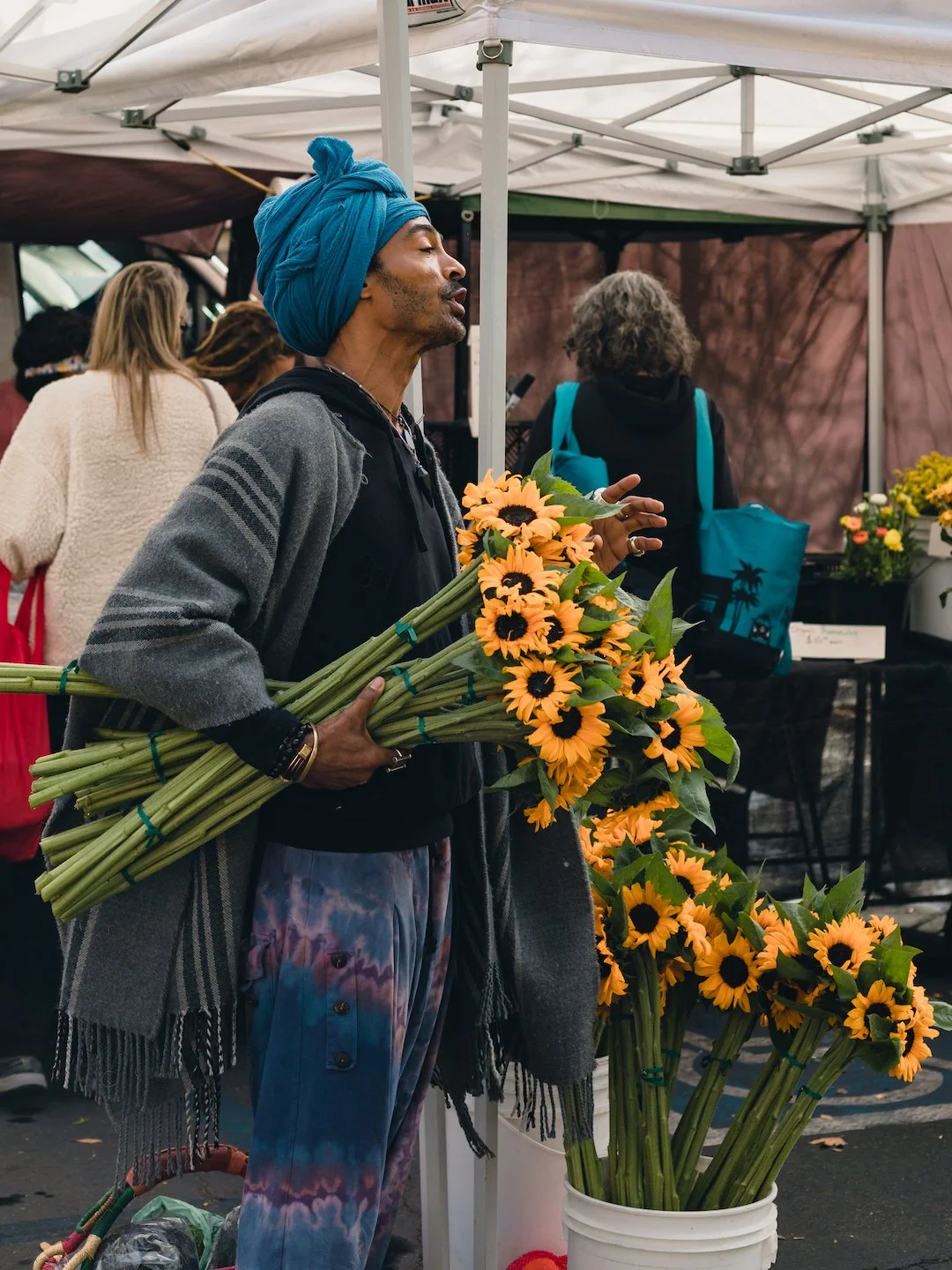 A man with a blue turban holding a large bouquet of sunflowers at an outdoor market.