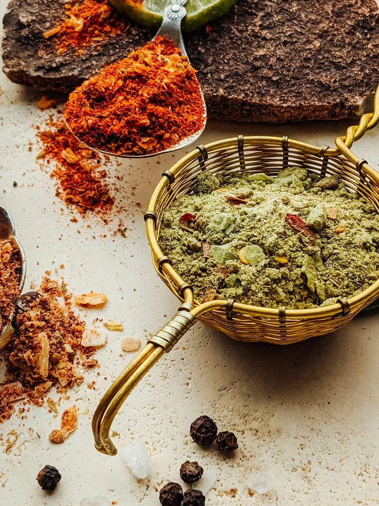 Close-up of various spices, including a greenish crushed herb in a small basket, red powder in a measuring spoon, and dark brown crushed spice, with a background of whole, dried herbs and peppercorns.