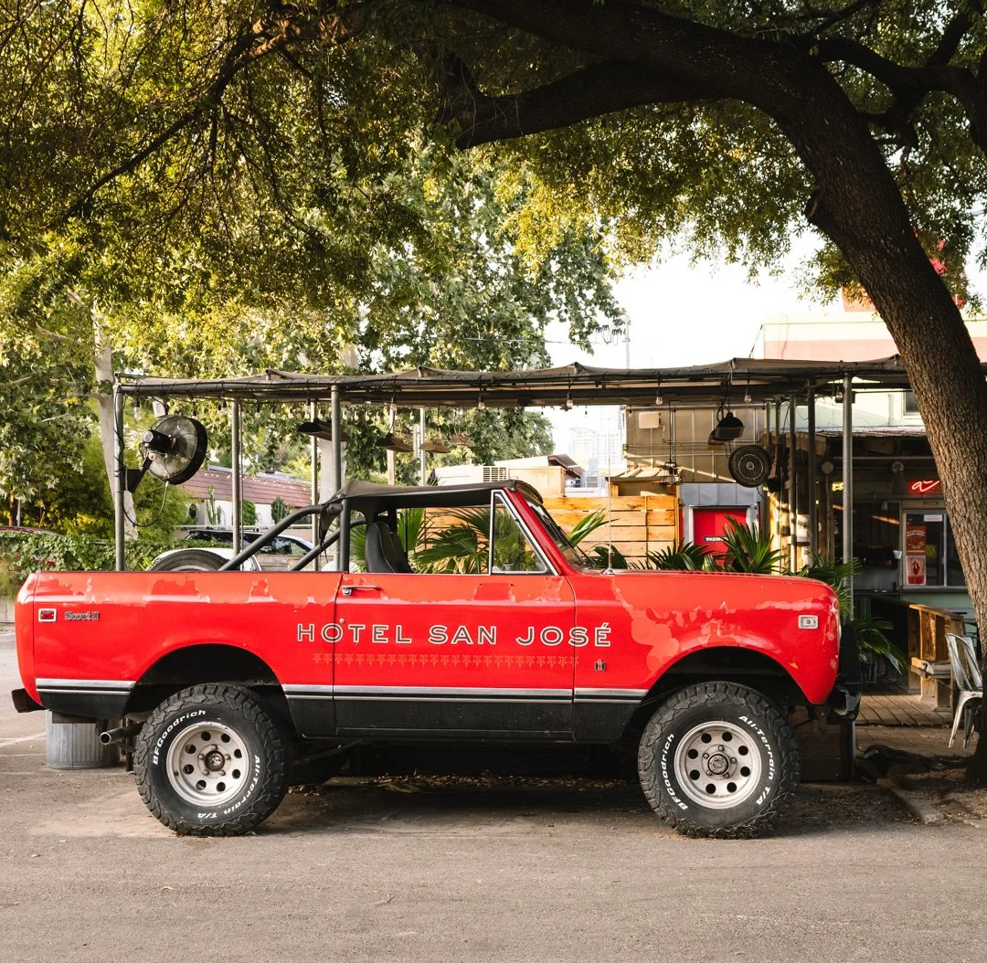 A red pickup truck with the words 'Hotel San José' written on the side parked on the street under a tree