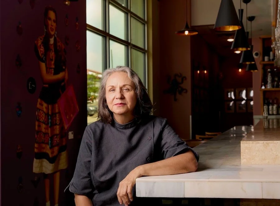 A middle-aged woman in a black chef's coat sitting at a marble bar counter inside a restaurant, with large windows behind her and modern pendant lights overhead.