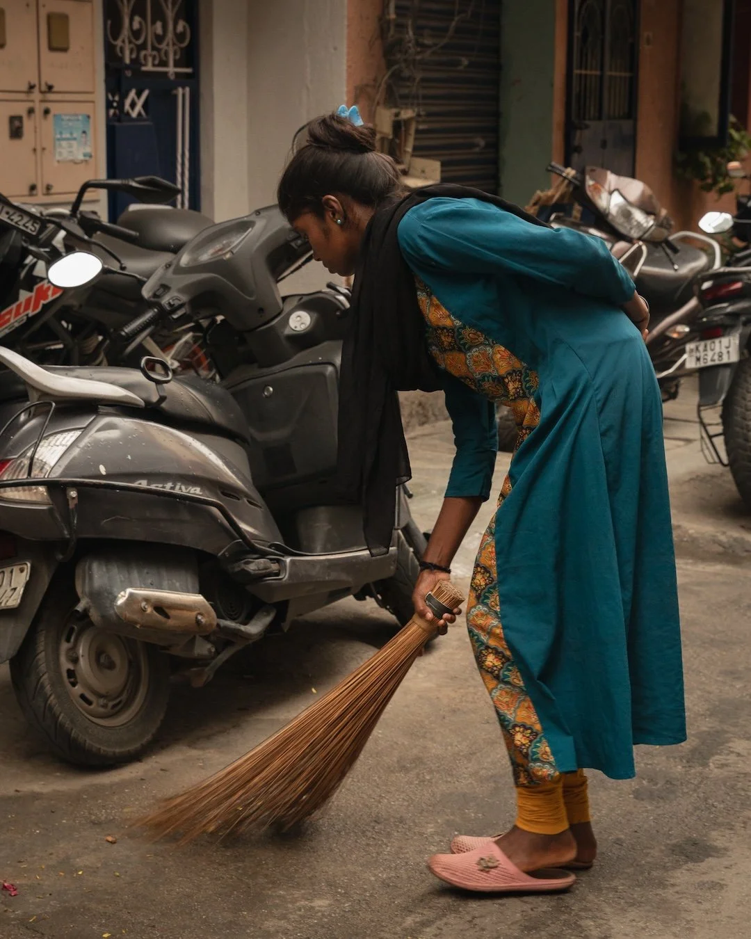 A woman in a blue traditional dress sweeping the street with a broom near parked motorcycles.
