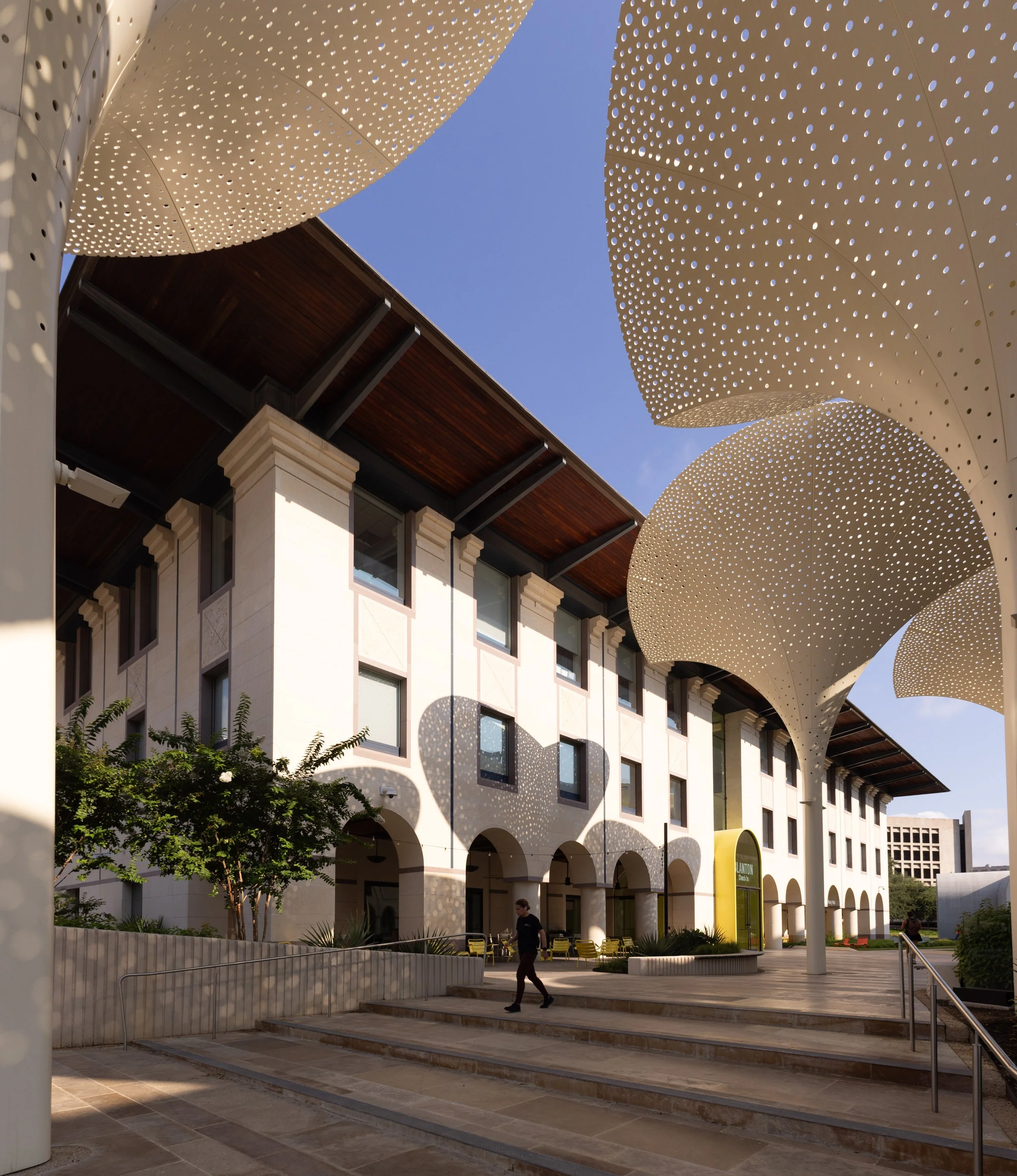 A modern building with perforated, wave-shaped white metal panels overhead, providing shade. The building has large windows and columns, with a landscaped area in front. People are walking, and there are yellow chairs on the patio.