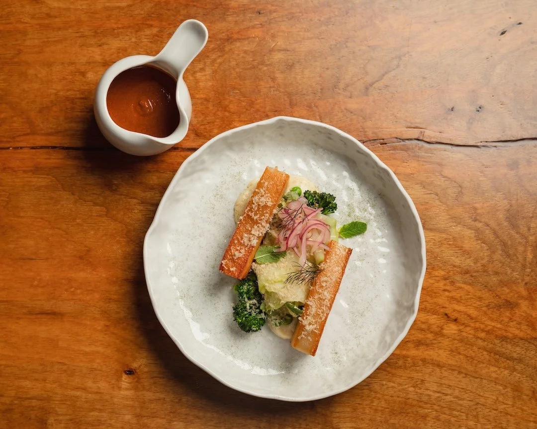Plate with two toasted bread sticks, broccoli, greens, and sliced radishes, with a small pitcher of sauce on the side.