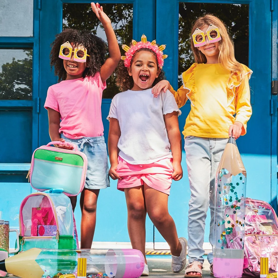 Three young girls celebrating outdoors with colorful backpacks and party props, smiling and making joyful gestures.