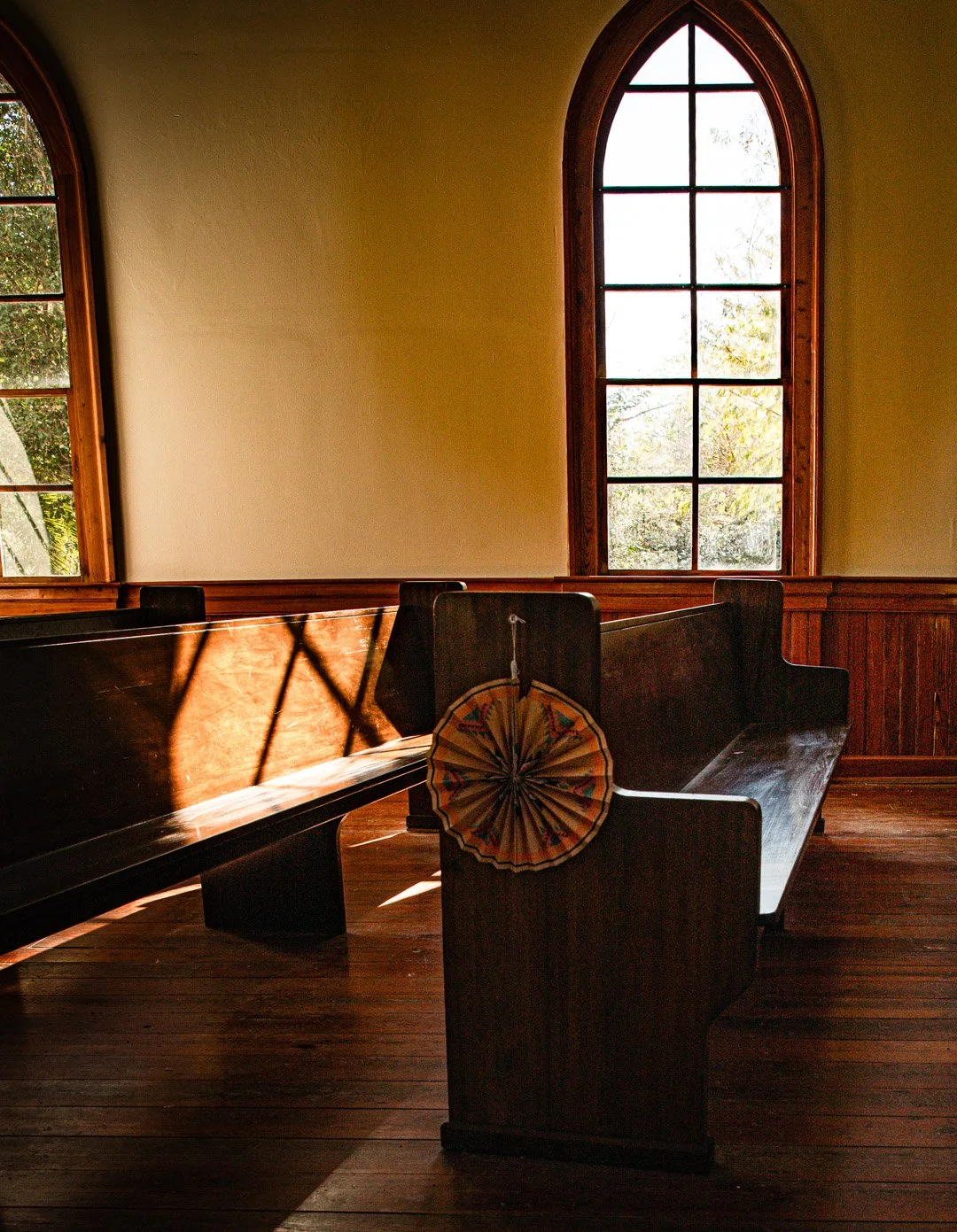 Interior of a church with wooden pews, tall arched windows, and sunlight streaming in, creating shadows on the pews, with a decorative fan hanging on one pew.